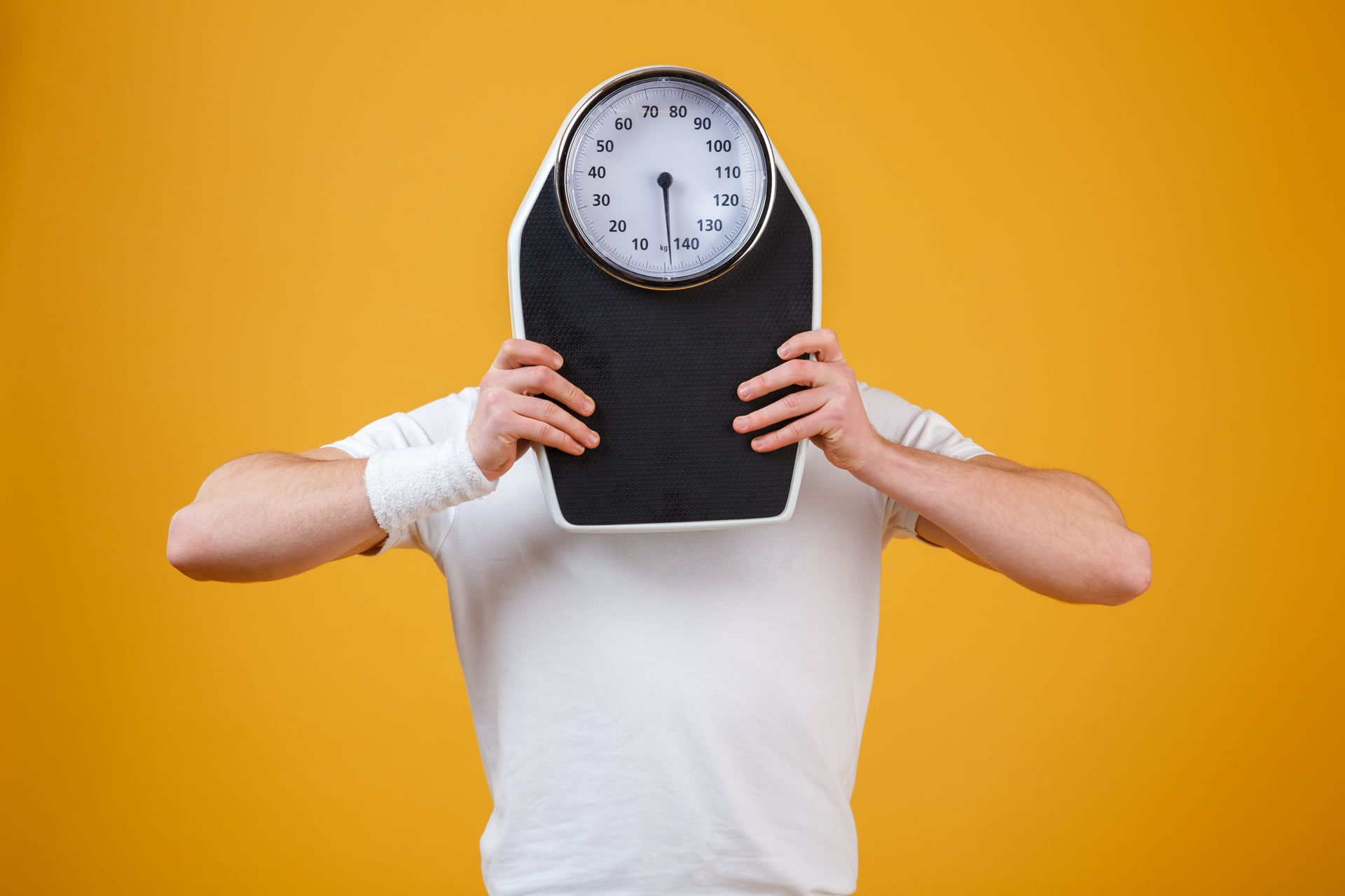 Young sports man hiding face behind weight scales